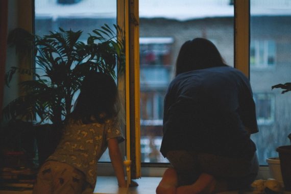 Two figures, possibly a mother and child, quietly look out of a window surrounded by indoor plants, observing the world outside.