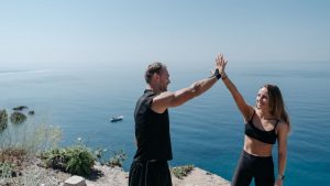 A happy couple in activewear high fives on a cliff by the ocean, enjoying a sunny day outdoors.